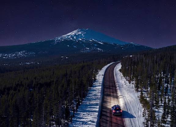 A car is driving down a snowy road with a mountain in the background.