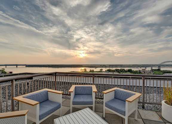 Two chairs and a table on a balcony overlooking a river at sunset.