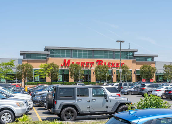 The bustling Market Basket parking lot in Chelsea, MA, with a large grocery store and multiple cars parked outside under a clear blue sky.