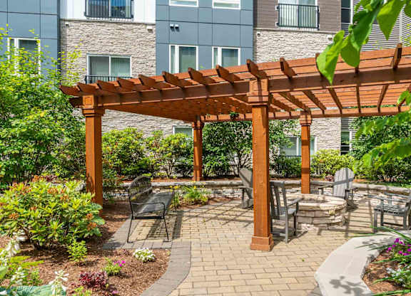 The Residences at Crosspoint central courtyard at 42 Wellman Street in Lowell, MA features paved walkways, raised garden beds, green shrubs, benches, and surrounding multi-story buildings clad in stone and metal siding.