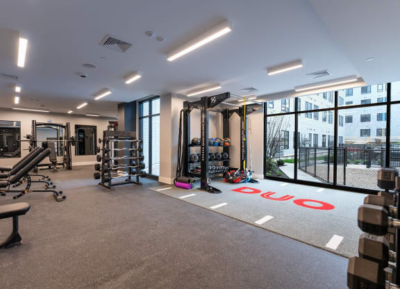 The wide, open space of the DUO Apartments fitness center, featuring racks of weights, benches, TRX suspension systems, and a functional training area with large windows letting in natural light.