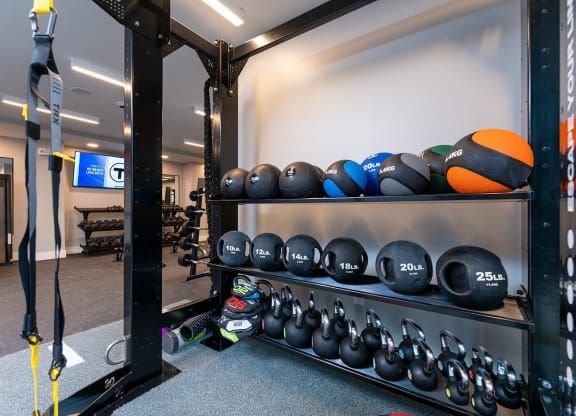 A fitness area at DUO Apartments featuring TRX suspension trainers, kettlebells, and medicine balls neatly organized on racks, with a spacious workout area and weightlifting benches in the background.