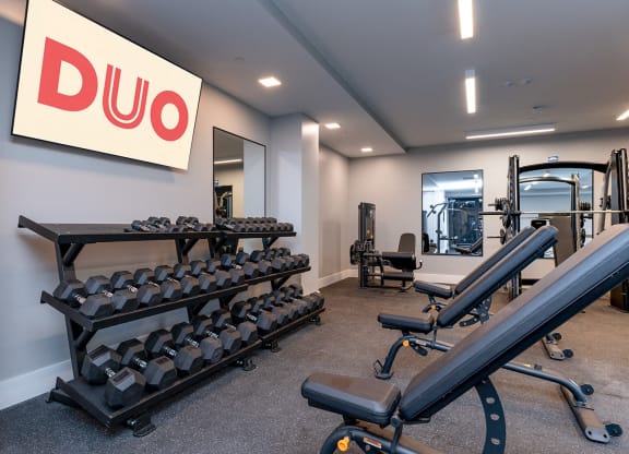 DUO Apartments gym featuring multiple adjustable benches, dumbbell racks, and a DUO-branded screen on the wall, creating a modern and organized workout space.