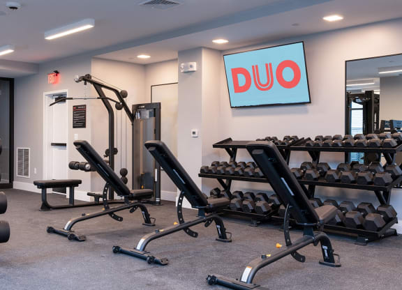 DUO Apartments' fitness center, featuring weightlifting equipment, benches, and a well-lit, spacious area for workout routines.