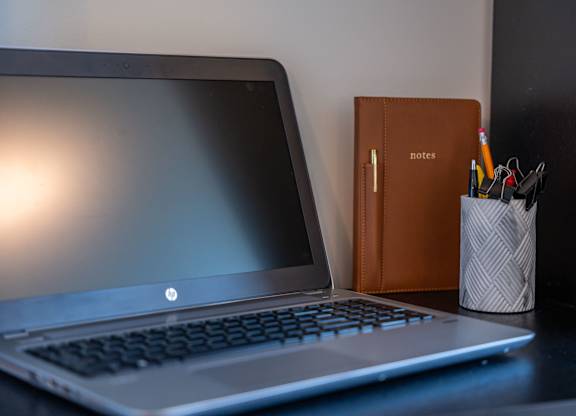 A laptop with a notebook and a cup of pens on the side on a desk at the Mastlight.