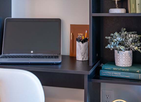 A laptop is on a desk with a white chair, a pencil holder, and a book titled "Brightwood" on a shelf at the Mastlight.