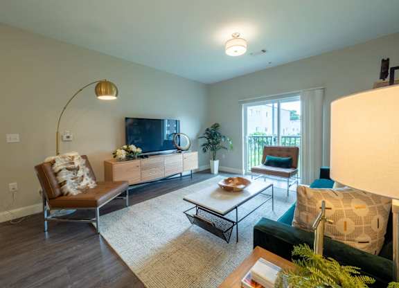 A bright living room with floor-to-ceiling sliding doors leading to a private balcony, furnished with modern seating and a flat-screen TV.