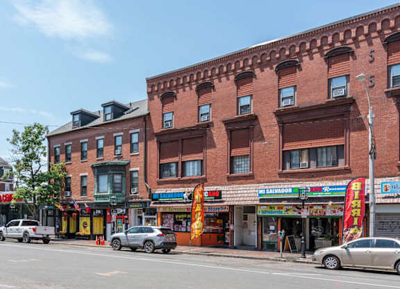 A street view of a city with cars parked and buildings with shops on the ground floor.