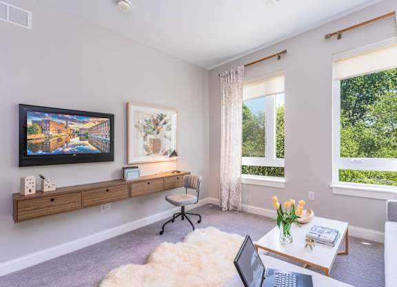The Residences at Crosspoint living room corner features a wall-mounted flat-screen TV, abstract artwork above a floating wood console, a sheepskin rug on the floor, and a white coffee table holding a vase of yellow tulips.