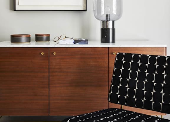 A black and white patterned chair sits in front of a wooden cabinet with a framed picture above it.