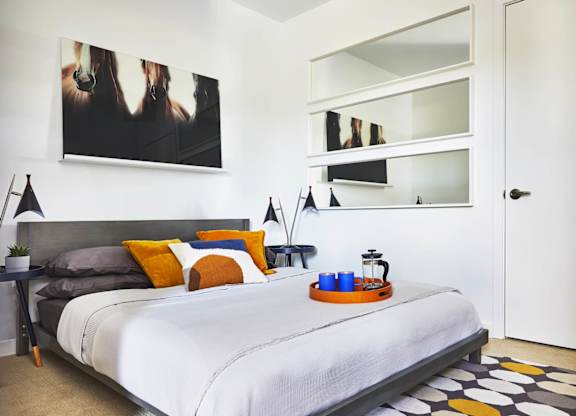 A bedroom with a bed, a nightstand with a tray on it, and a painting on the wall, model apartment