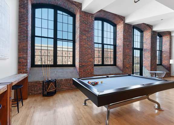 A pool table is in the middle of a room with brick walls and large windows.
