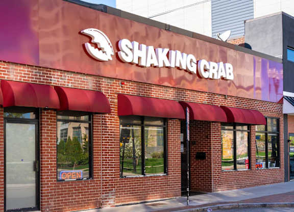 The exterior of a Shaking Crab restaurant with a red awning and a blue sign on the window.