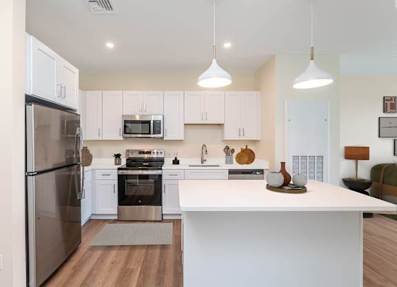 Staged kitchen featuring white cabinetry, stainless appliances and a breakfast island topped with decorative pottery under two modern pendants.