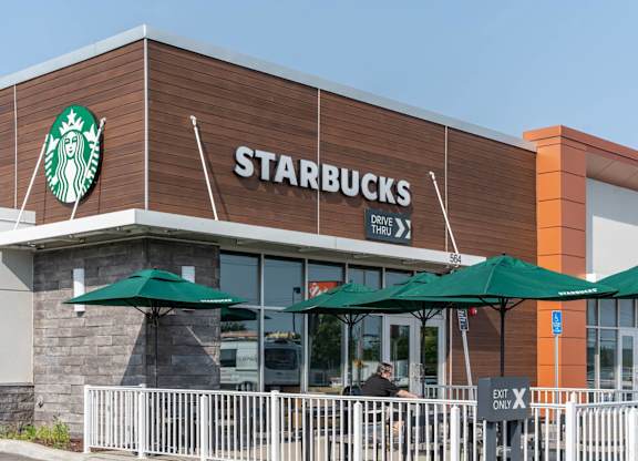 Starbucks café with wood-accent façade, drive-thru sign and patio tables shaded by green umbrellas.