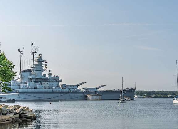 Historic battleship anchored on a calm river alongside small sailboats and tree-lined shoreline.