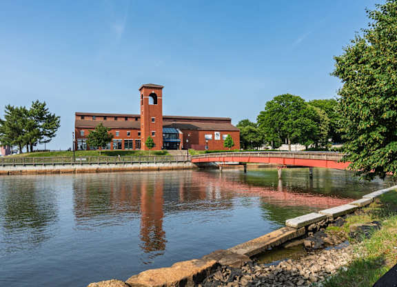 Brick waterfront museum with tower reflected in the river, connected by a red pedestrian bridge beneath a clear sky.