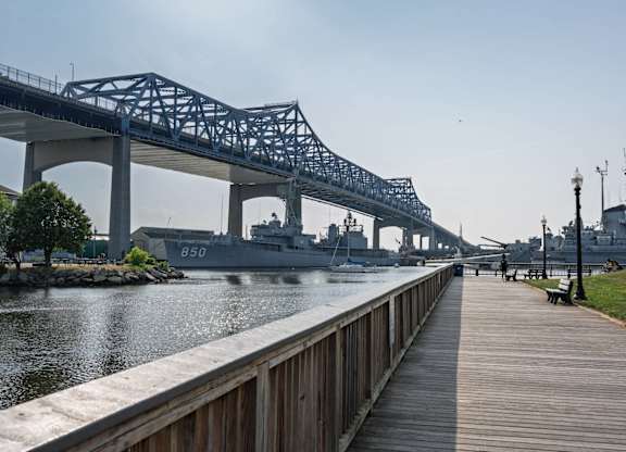 Boardwalk beneath a steel truss bridge, looking toward moored naval ships along the sparkling river.