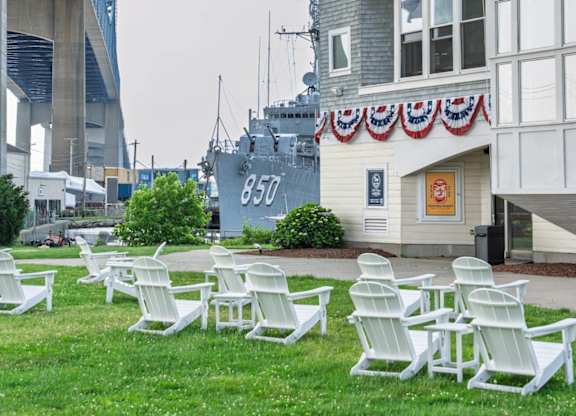 Lawn with white Adirondack chairs beside a carousel pavilion draped in patriotic bunting, with destroyer 850 and bridge backdrop at Battleship Cove