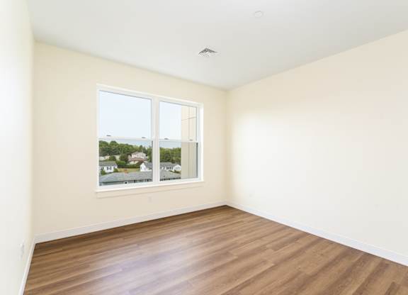 Sun-filled, empty bedroom showcasing natural-tone plank floors and a wide picture window.