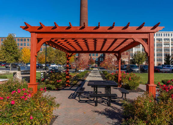 a picnic table with a fire pit and grills under a wooden pavilion on a brick sidewalk