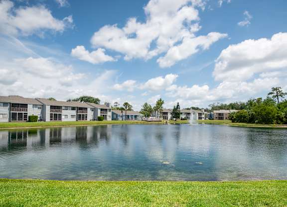 A serene lake in front of a row of houses under a blue sky with clouds Fishermans Landing Apartments in Ormond Beach, FL.