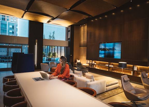 A woman in an orange shirt is sitting at a long white table in a room with a large screen on the wall.
