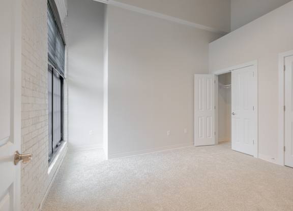 A room with white walls and carpeted floor with a window on the left and three doors on the right.