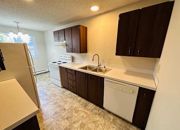 Galley-style kitchen with white appliances, dark wood cabinets, laminate countertops, and vinyl flooring in 1-bedroom Bridgeport Terrace apartment