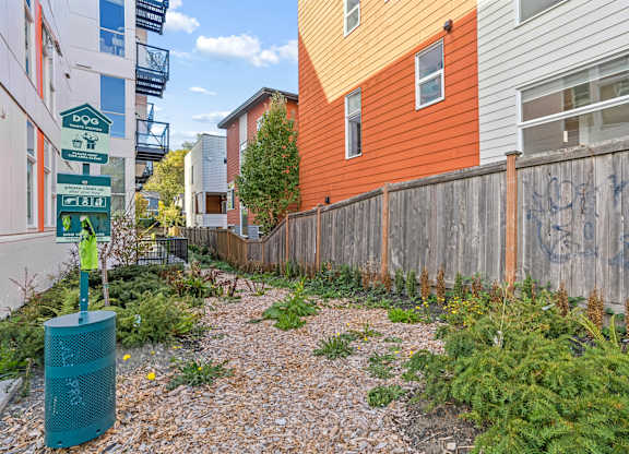 A gravel path leads to a green trash can in a narrow alley between two buildings.