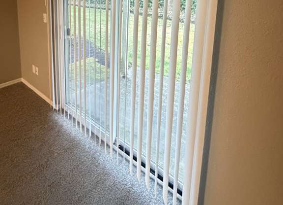 A white vertical blind is open to a view of a green lawn.