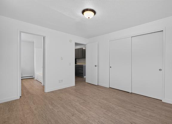 A room with white walls and wooden flooring with a refrigerator and cupboards in the background.