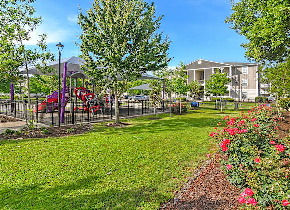 A playground with a red slide and a purple canopy is surrounded by a green lawn and a fence at Gulf Hills Apartment Homes in MS 39564
