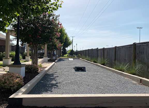 A long, narrow, gravel-covered pathway stretches into the distance between a wooden fence and a row of trees.
