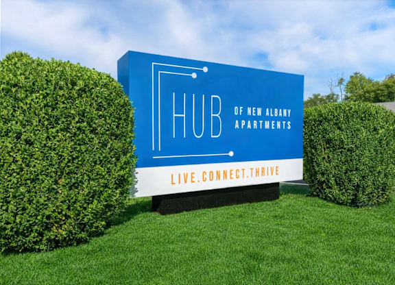 A blue sign for Hub of New Albany Apartments sits in front of two bushes.