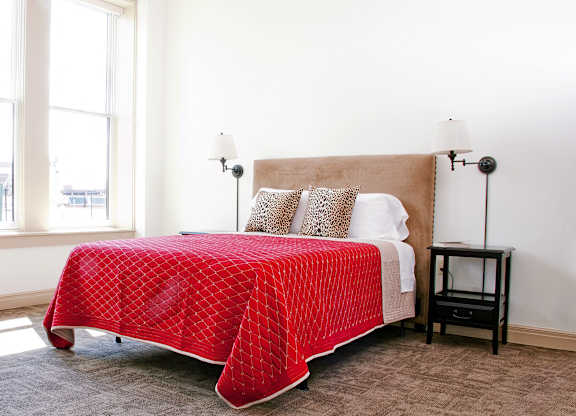 a bedroom with a bed with a red blanket and two windows at 100 South Apartments, Buffalo, New York