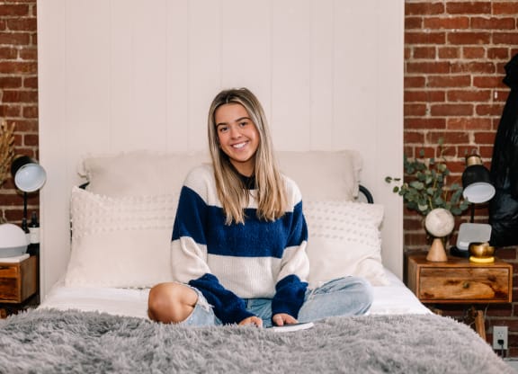 a woman sitting on a bed with a brick wall behind her at University Commons Apartments, Oakland, Pittsburgh