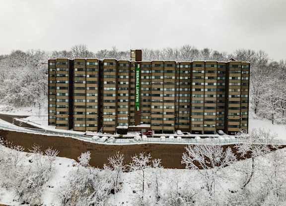 A tall building with a green sign on the side stands in front of a snowy landscape at Walnut Towers at Frick Park, Pittsburgh, PA