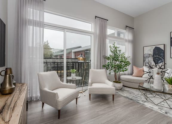 2-Bedroom Apartments in Liberty Township, OH - Liberty Center - Furnished Living Room with Vinyl Wood Plank Flooring, Area Rug, Sofa, Coffee Table, Armchairs, Houseplant, Storage Cabinet, Wall-Mounted TV, Large Window, and Sliding Glass Door with Access to Balcony.