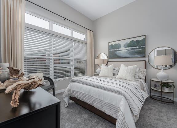 One-bedroom Apartments in West Chester OH - Liberty Center - Furnished Bedroom with Grey Shaw Carpeting, Round Coffee Tables with Lamps, Circular FRamed Mirrors, Chair, Wooden Dresser, Window