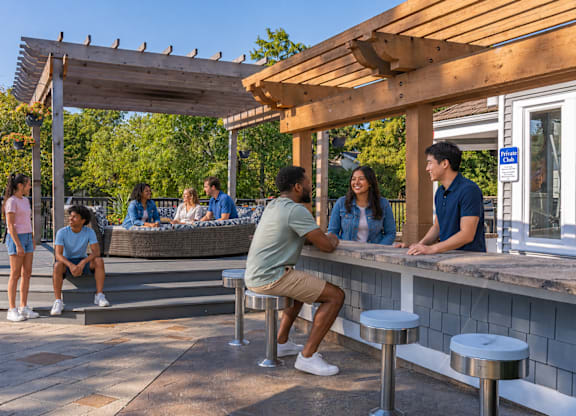A group of people are sitting and standing around a picnic table under a wooden pergola.