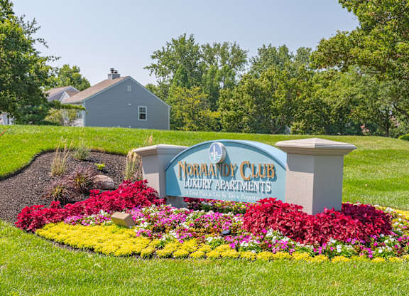A sign for Normandy Club Luxury Apartments is surrounded by flowers.