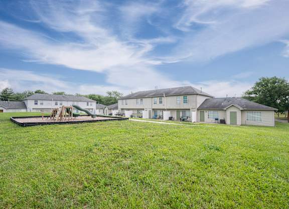 A large grassy field with a playground and houses in the background.