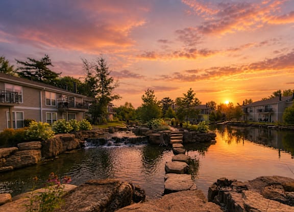 A serene landscape with a sunset over a pond with rocks in the foreground and houses in the background.