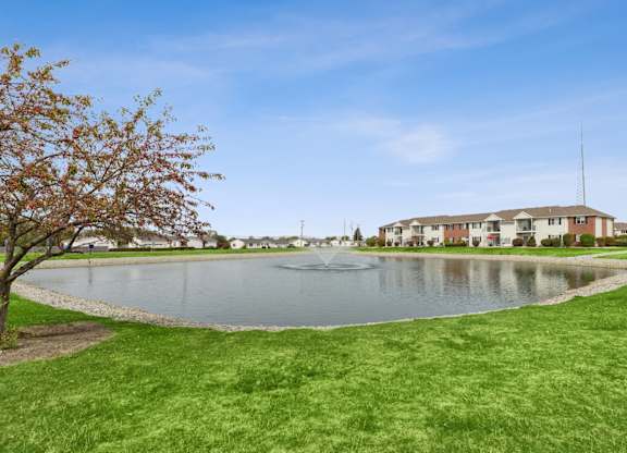 A tree with red leaves is in the foreground of a grassy area with a lake and a building in the background.