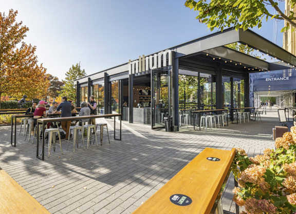 A group of people are sitting at tables under a glass and wood structure.