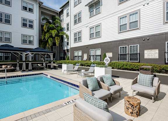 A pool surrounded by chairs and cushions with a building in the background.
