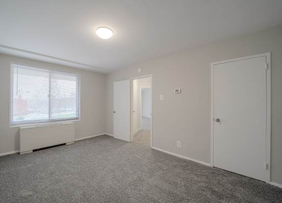 A room with a carpeted floor, a window with blinds, and a ceiling light.