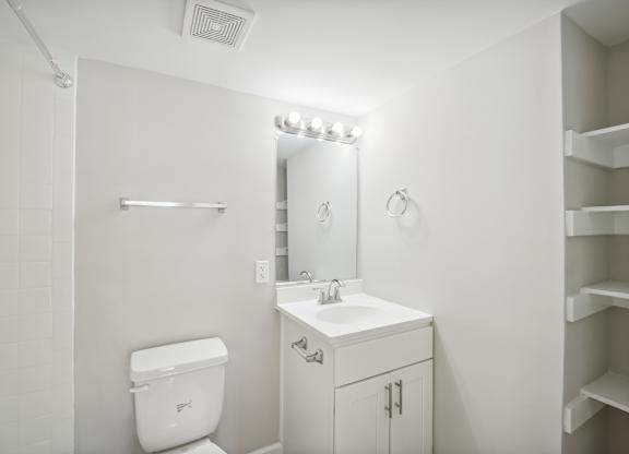 A white bathroom with a toilet, sink, mirror, and shelves.