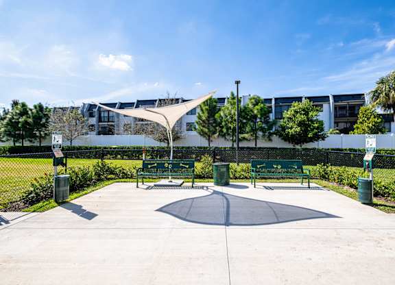 A playground with a round platform and a shade structure is surrounded by green bushes and trees.